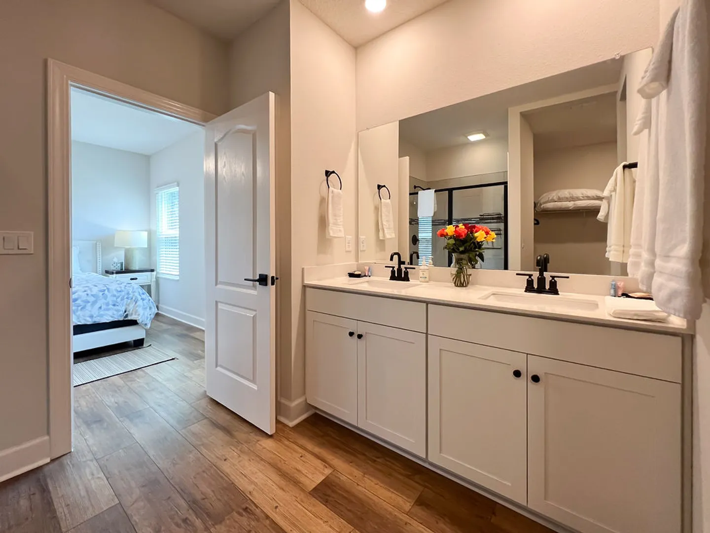 Bathroom with double vanity, large mirror, and view into the adjoining bedroom through an open doorway.