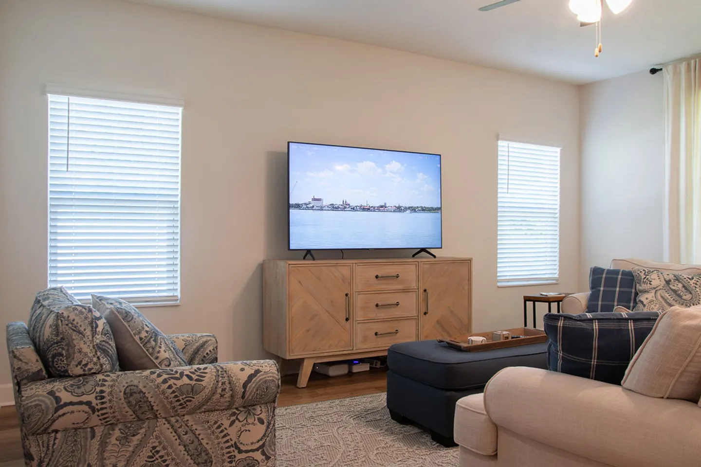 Living room with a large television on a light wood console table flanked by two windows with blinds, surrounded by patterned armchairs and an ottoman.