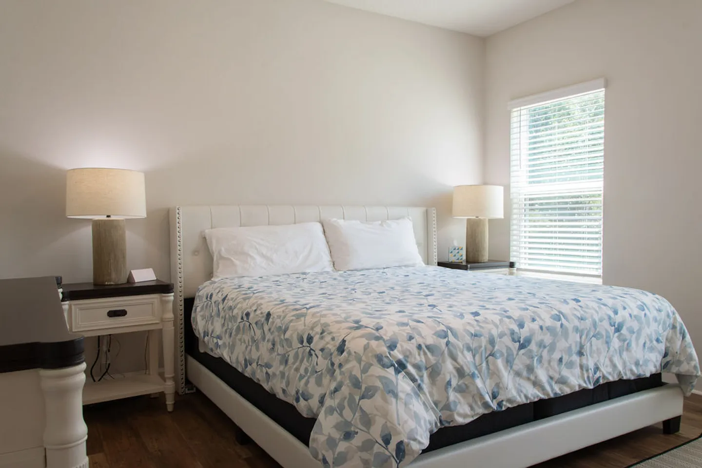 Bedroom featuring a bed with a white upholstered headboard, blue and white leaf-patterned bedding, two nightstands with matching lamps, and a window with blinds.
