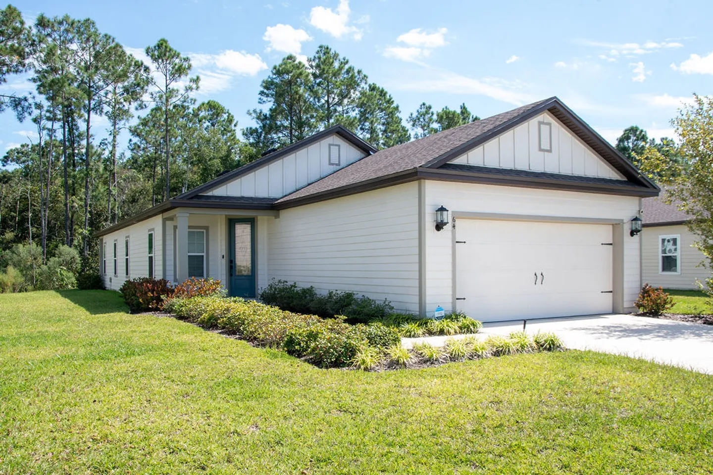 Exterior view of a single-story house with light siding, a two-car garage, manicured lawn, and pine trees in the background.