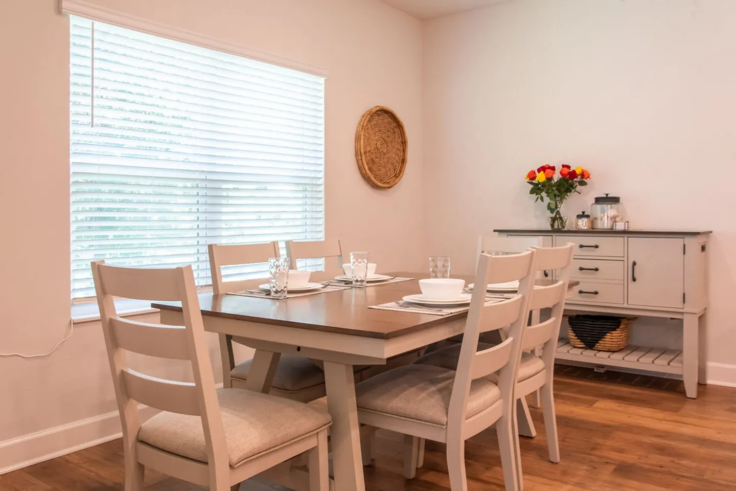 Dining area featuring a wooden table set with plates and glasses, surrounded by light-colored chairs next to a window with horizontal blinds.
