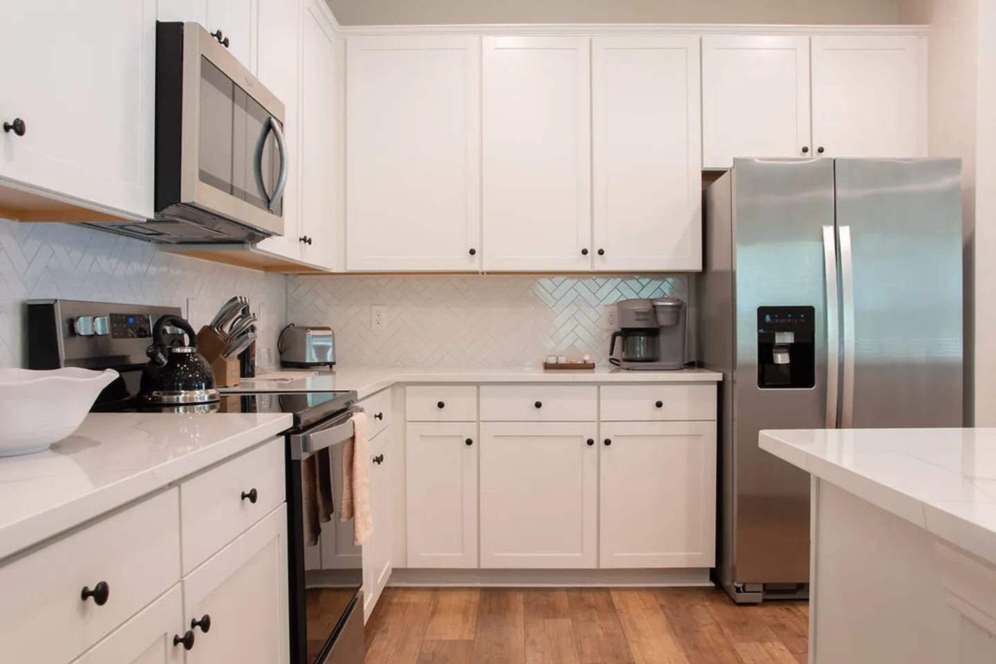 A kitchen with white shaker-style cabinets, light countertops, a herringbone tile backsplash, and stainless steel appliances including a refrigerator, microwave, and stove.
