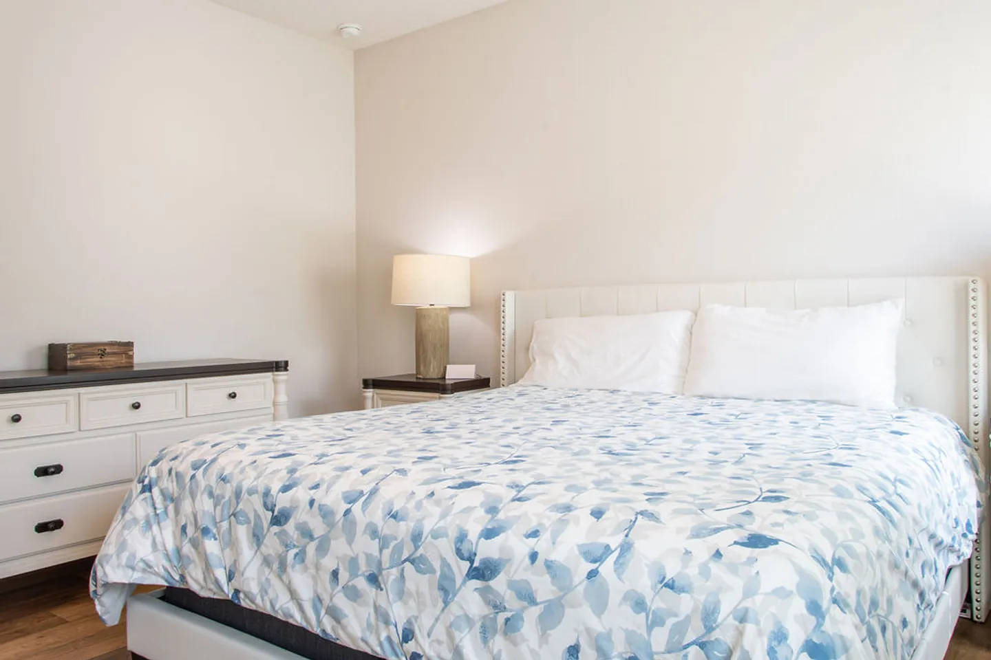 Bedroom with a white tufted headboard, blue and white leaf-patterned bedding, a nightstand with a lamp, and a white dresser.