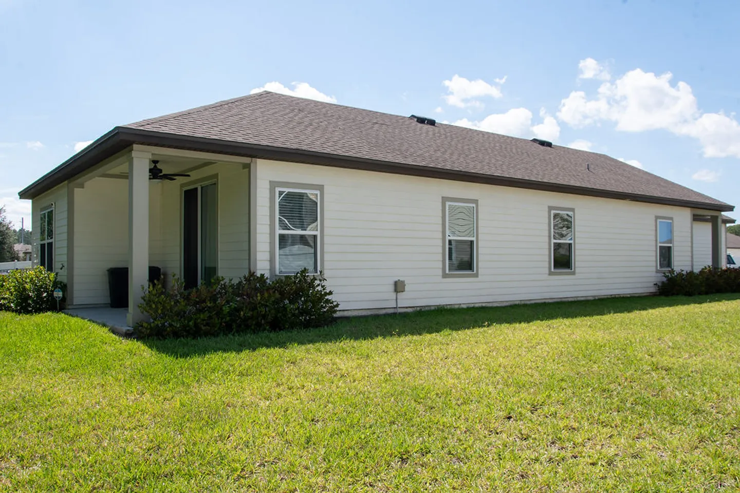 Exterior view of the back of a single-story home with light siding, a covered patio, and a grassy lawn under a partly cloudy sky.