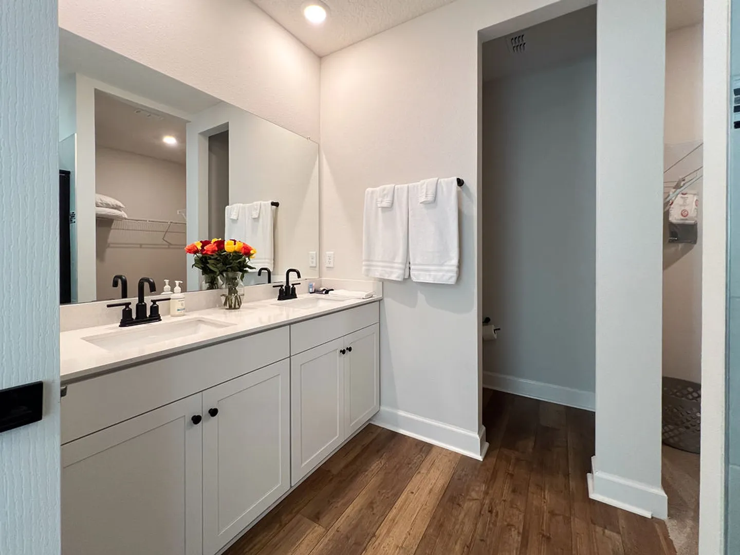 Bathroom featuring a double vanity with white cabinets, dark hardware, a large mirror, and wood-look flooring.