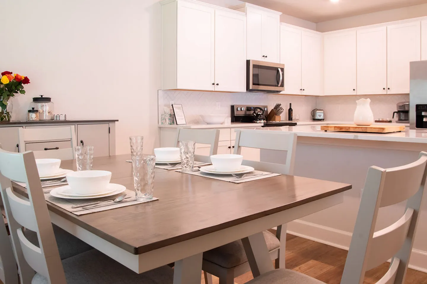 Dining table set with dishes and glassware in an open-concept kitchen with white cabinets.