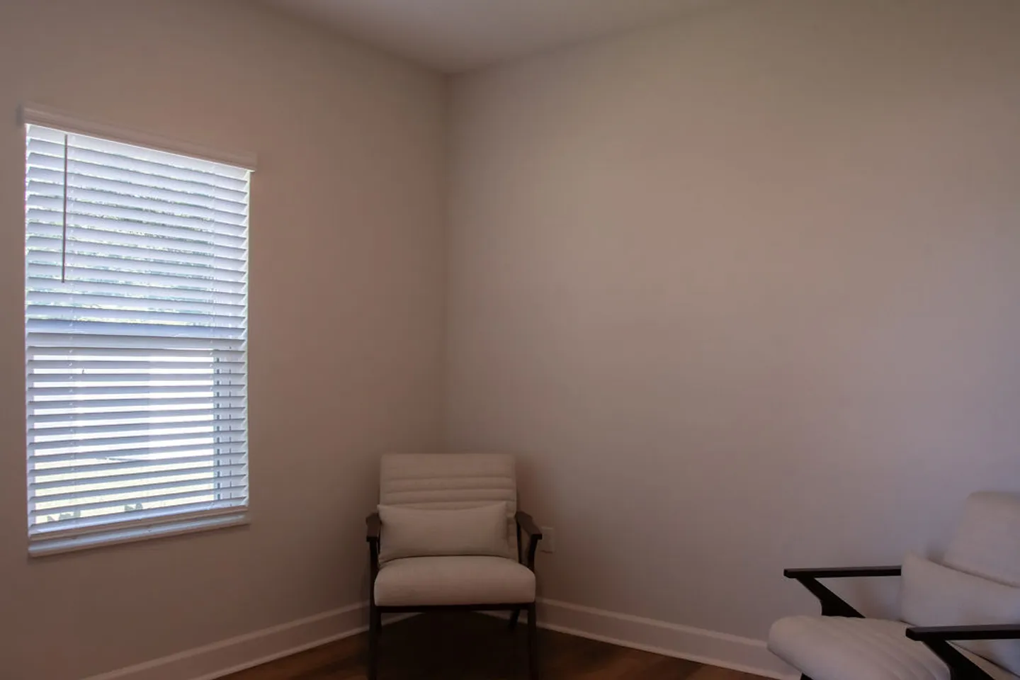 A corner of a room with light walls, wood floors, a window with horizontal blinds, and two armchairs.