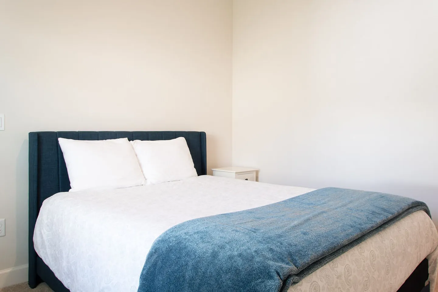 A made bed with an upholstered dark blue headboard, white linens, and a blue throw blanket set against plain white walls.