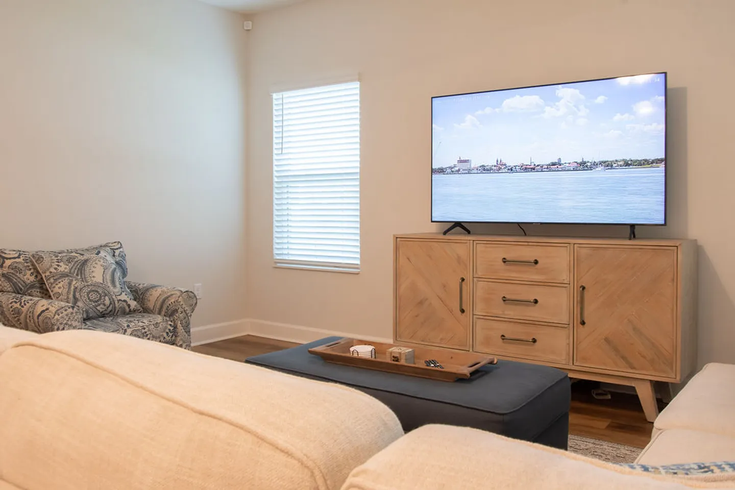 Living room area featuring a flat-screen television on a light wood console, viewed from behind a sofa with a cushioned ottoman and patterned armchair nearby.