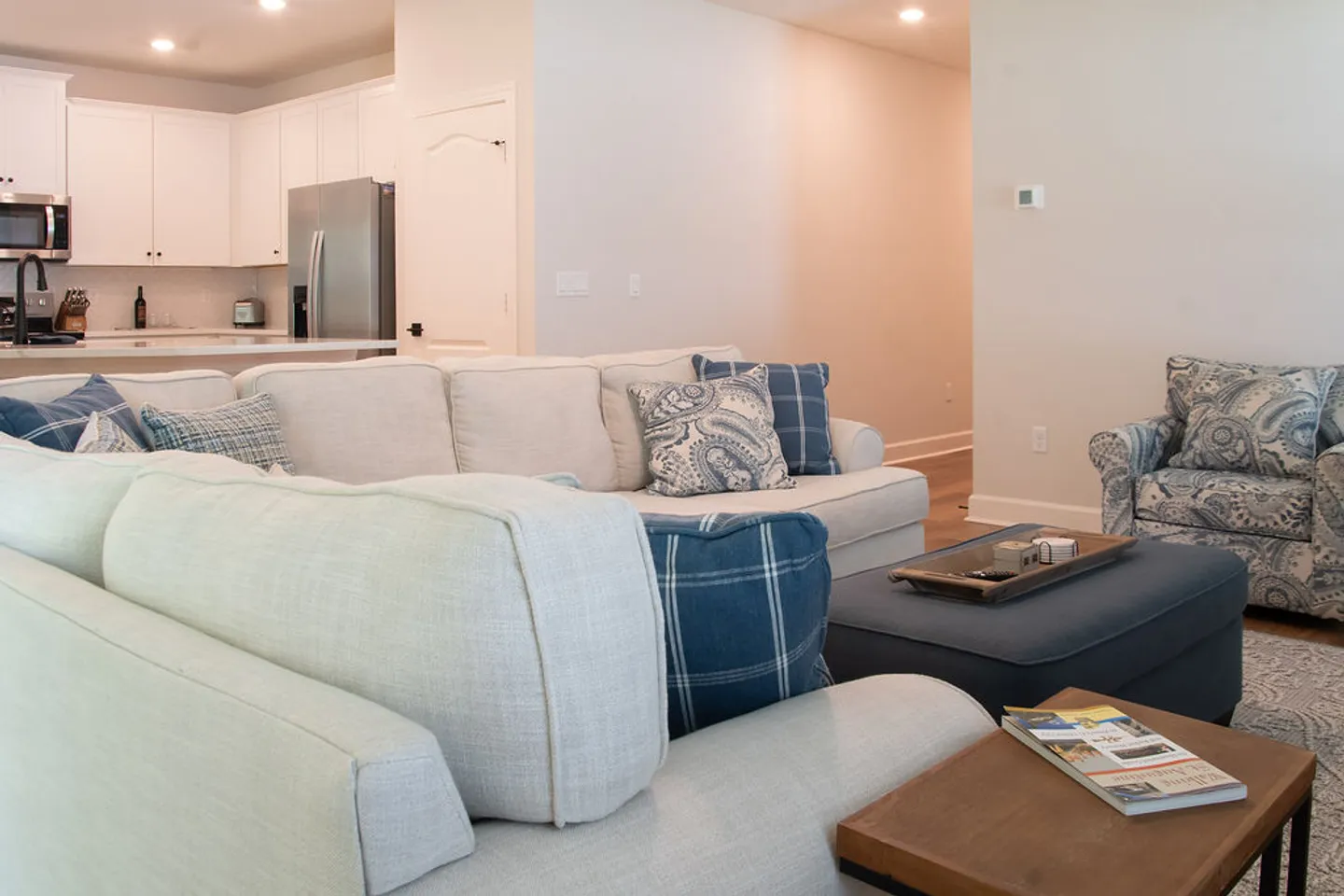 Living area with a light-colored sectional sofa, accent pillows, an ottoman coffee table, and patterned armchair, looking toward an open kitchen.
