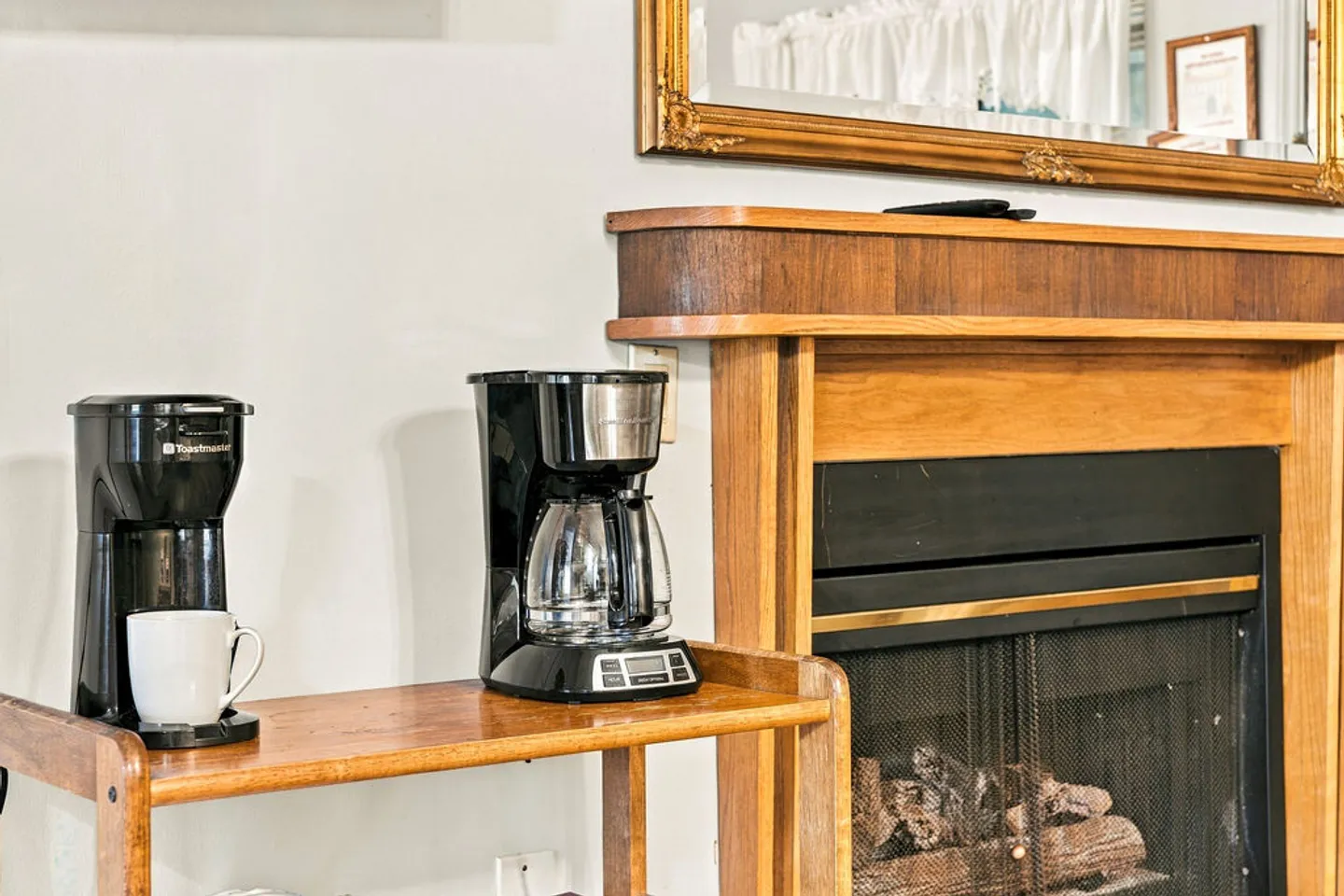 Two drip coffee makers on a wooden stand next to a fireplace with a wood mantel.