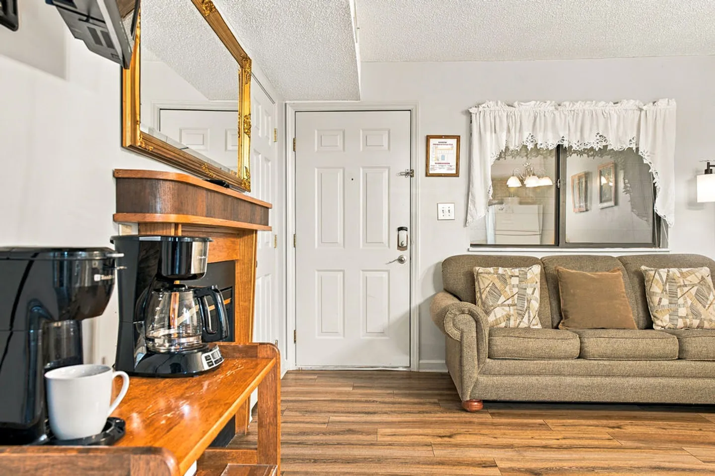 Living area showing an entrance door, a coffee maker on a wooden table, and an upholstered sofa next to an interior window.