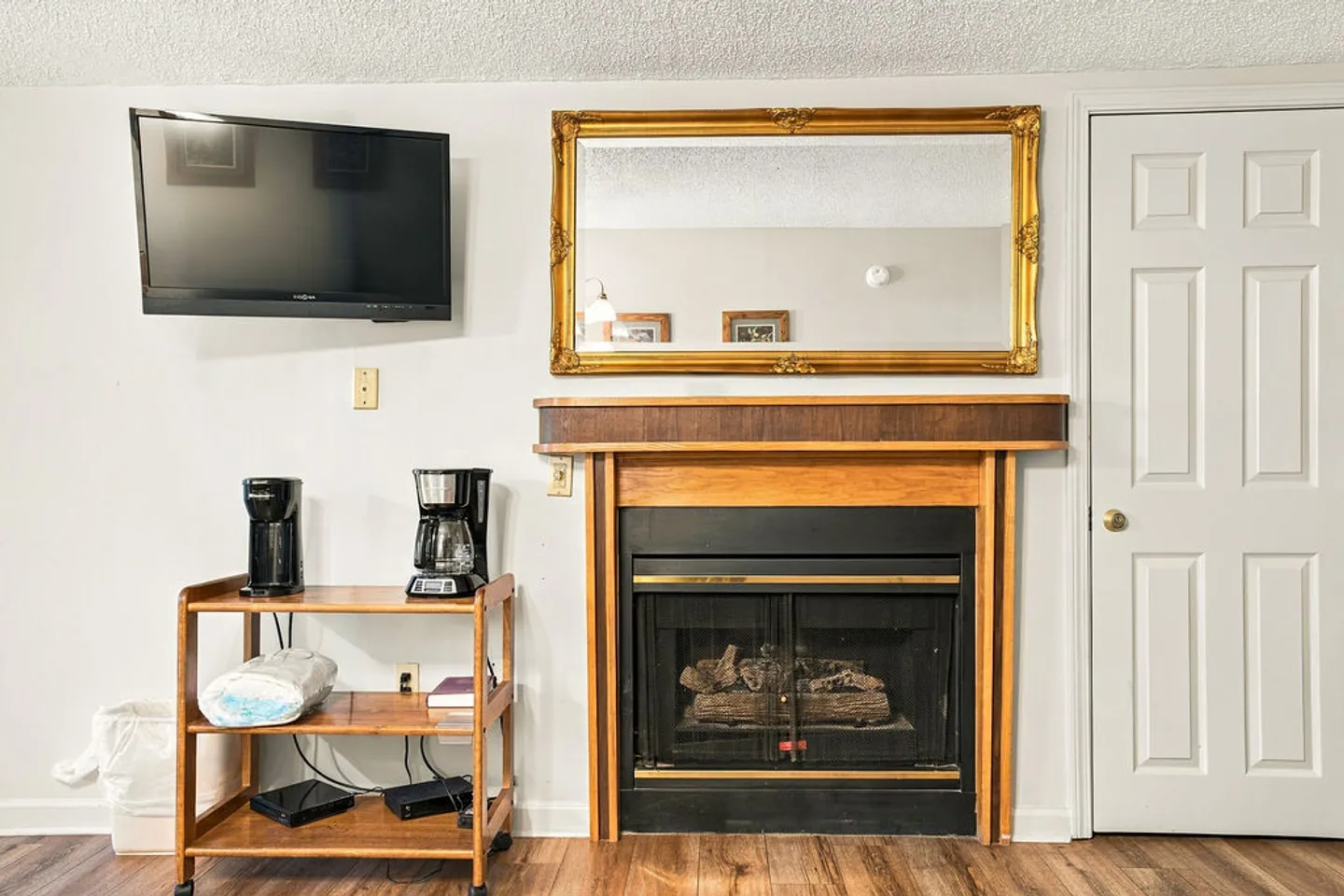 Living area featuring a fireplace with a wood mantel, a gold-framed mirror above, a wall-mounted TV, and a cart with coffee makers.