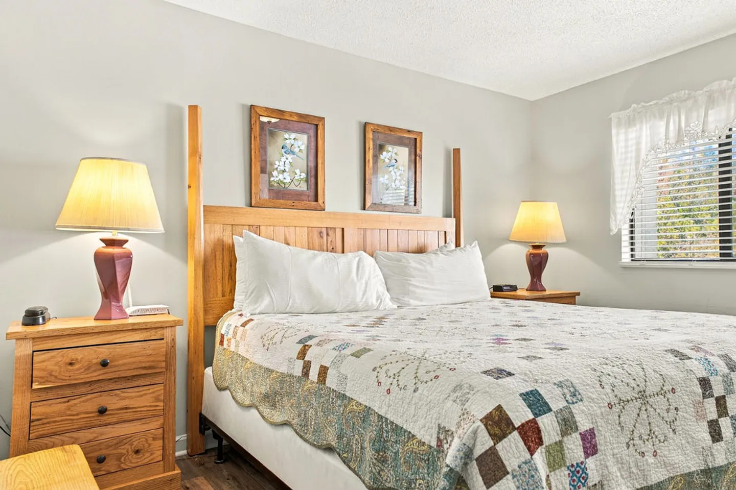 Bedroom with a wooden bed, patterned quilt, matching wooden nightstands with lamps, and a window with white curtains.