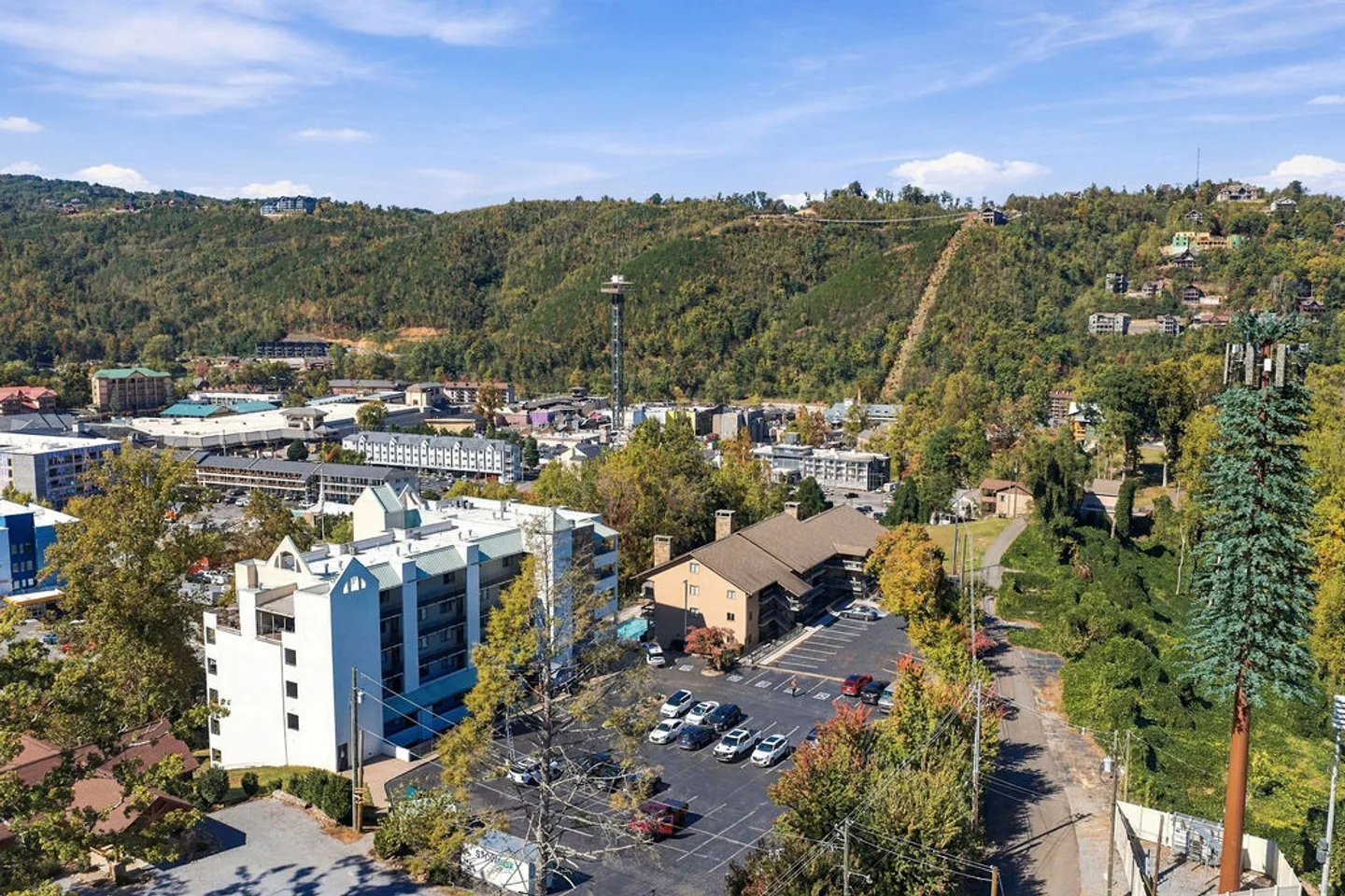 Aerial view of a multistory building complex and parking lot surrounded by trees, with a town and forested hills in the background.
