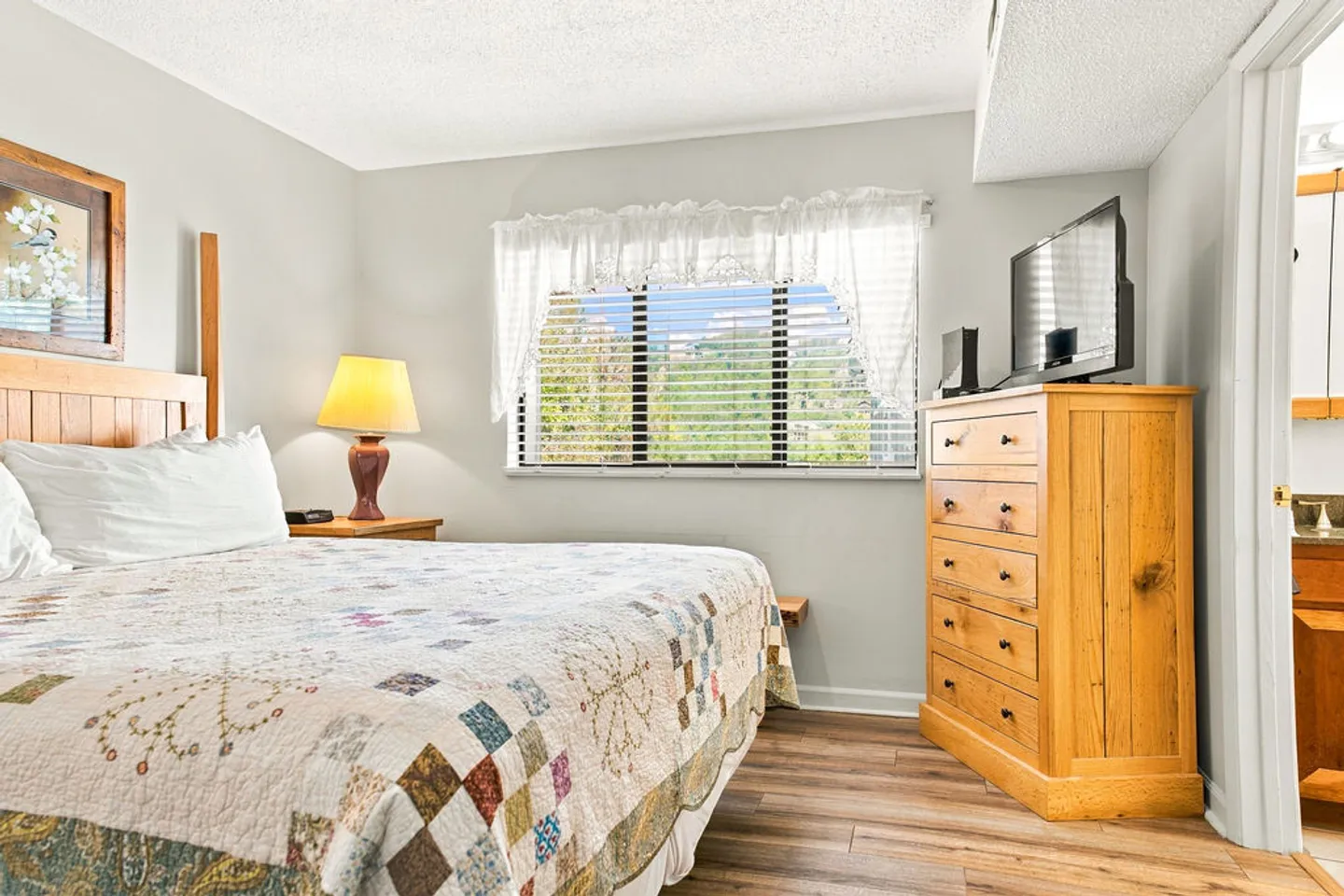 Bedroom with a wooden headboard, patchwork quilt, tall wooden dresser with television, and window with white valance curtains.