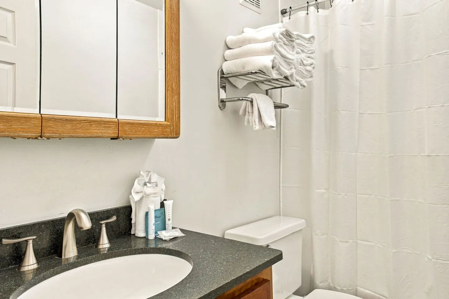 Bathroom showing a vanity with dark countertop, medicine cabinet, toilet, towel rack with folded towels, and a white shower curtain.