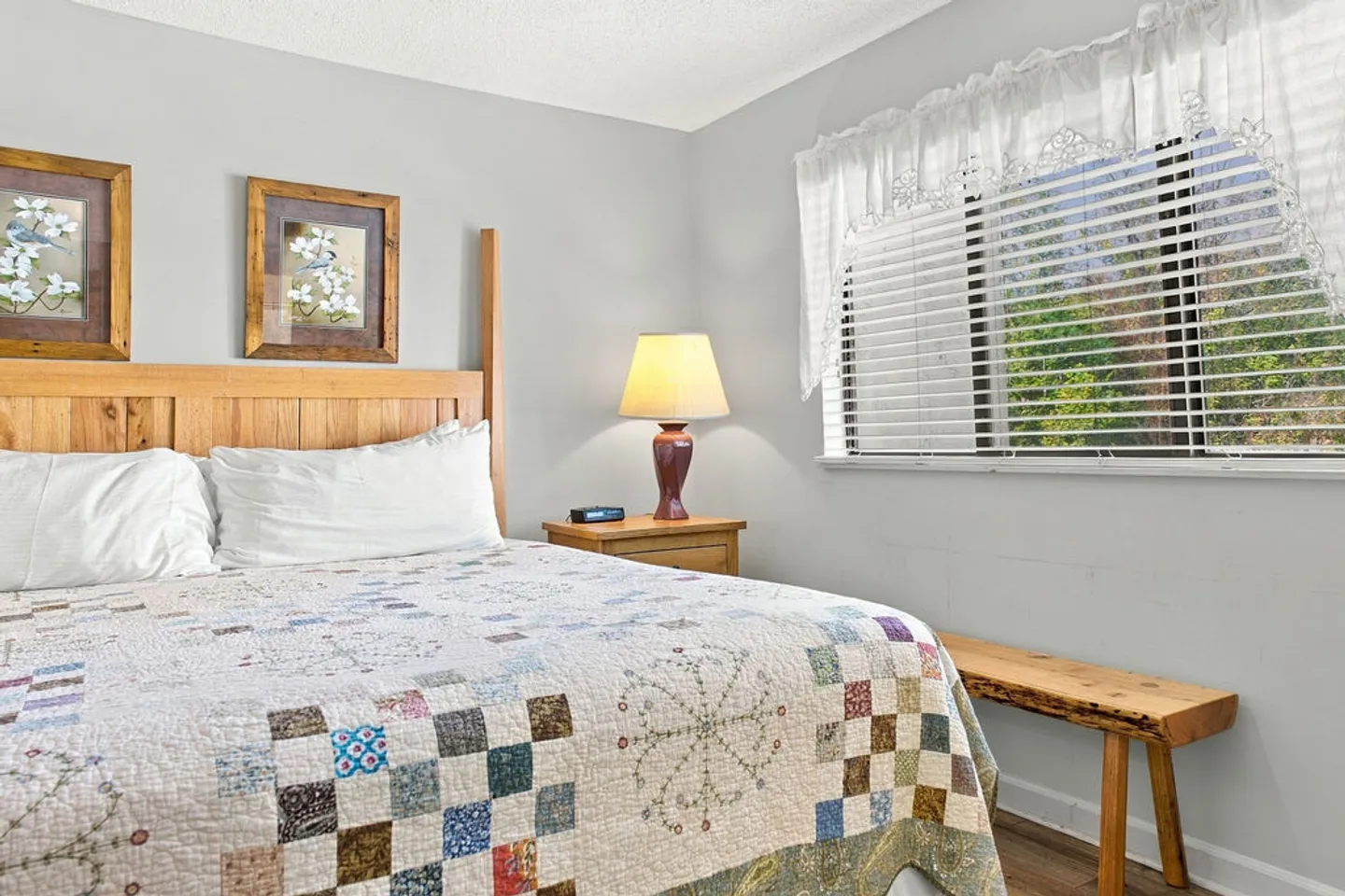 Bedroom with a wooden bed, patterned patchwork quilt, nightstand with lamp, and a window with blinds.