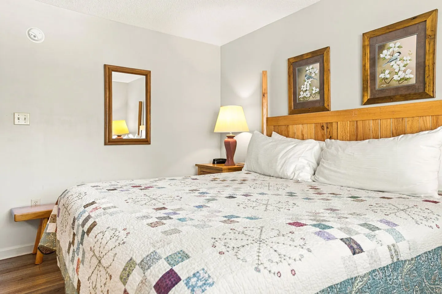 Bedroom with a wooden headboard, patchwork quilt on the bed, nightstand with a lamp, and framed artwork on the wall.