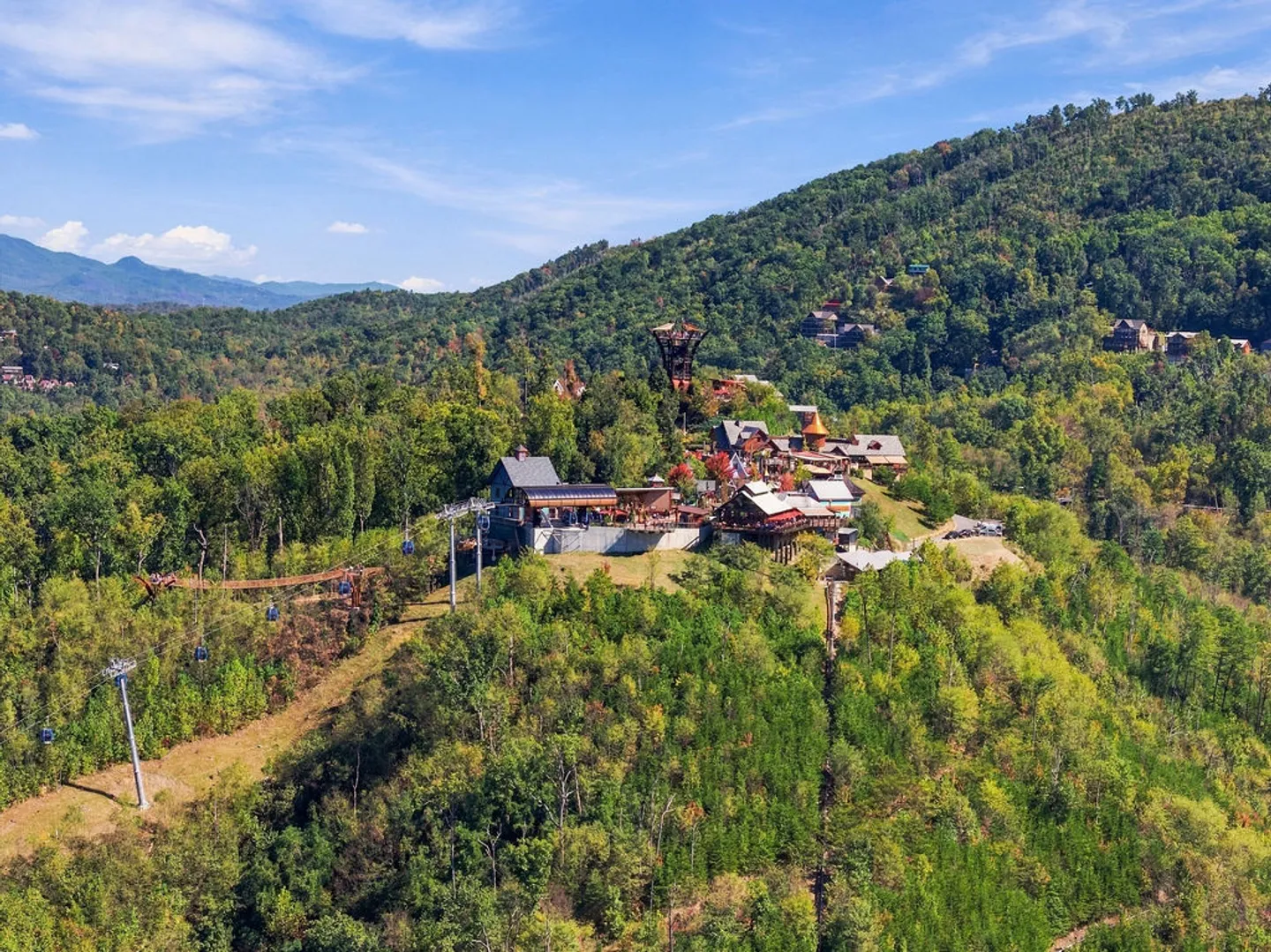 Aerial view of a mountaintop village attraction with chairlifts and suspension bridges surrounded by dense forest.