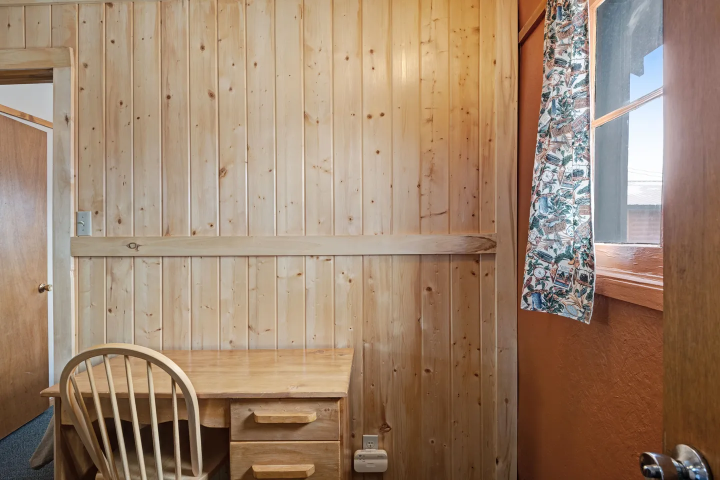 A small room corner featuring light wood-paneled walls, a matching wooden desk with a chair, and a window with patterned curtains.