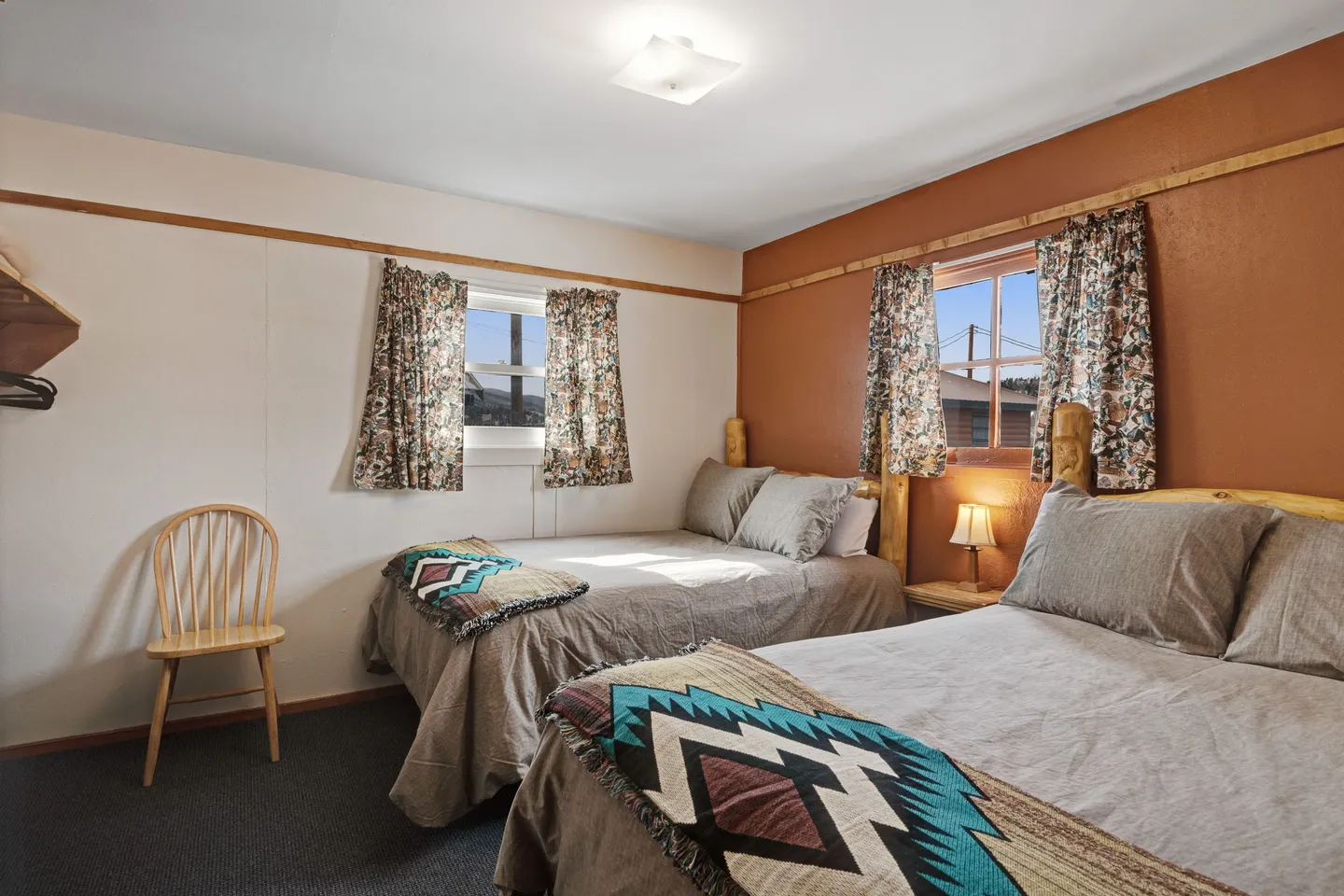 Bedroom featuring two beds with wooden log headboards, neutral linens, patterned throw blankets, and windows with floral curtains.