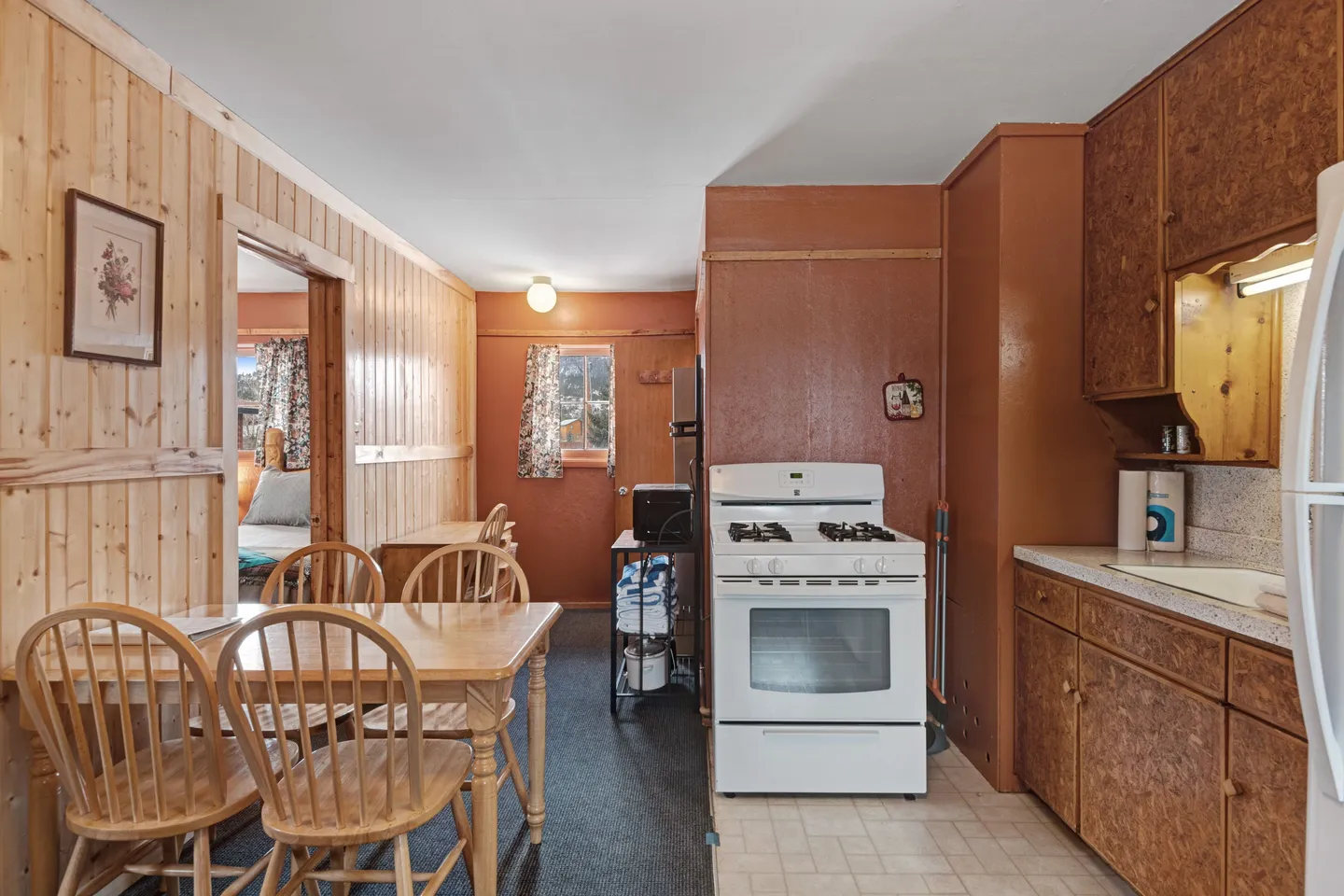 Kitchen and dining area featuring wood-paneled walls, a dining table with wooden chairs, white gas stove, and rustic cabinetry.