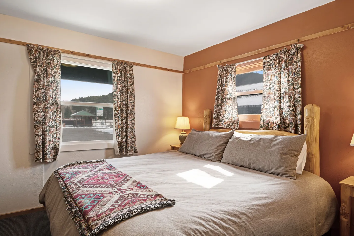 Bedroom with a log-style bed, neutral bedding with a patterned runner, and windows framed by floral curtains.