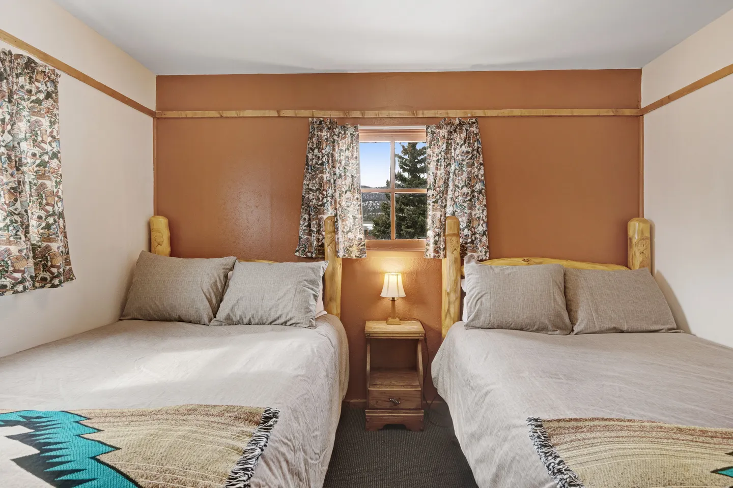 Bedroom featuring two beds with rustic wooden headboards and a nightstand under a small window with patterned curtains.
