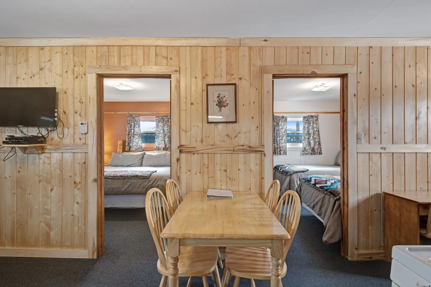 Dining table and chairs in front of a light wood-paneled wall with doorways leading into two bedrooms.