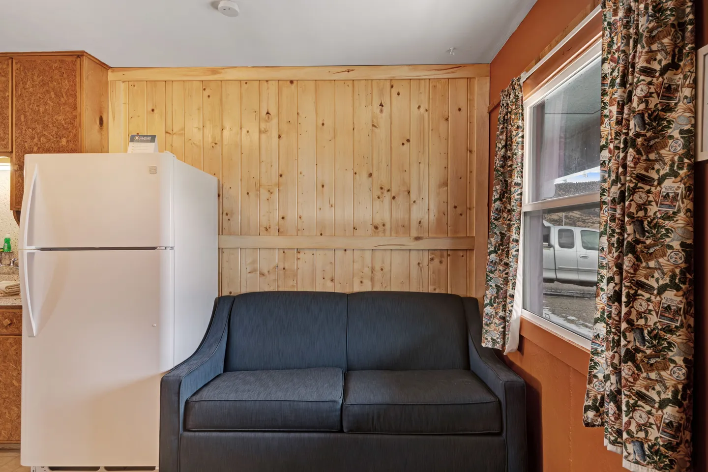 A dark fabric sofa placed against a wood-paneled wall next to a white refrigerator and a window with patterned curtains.
