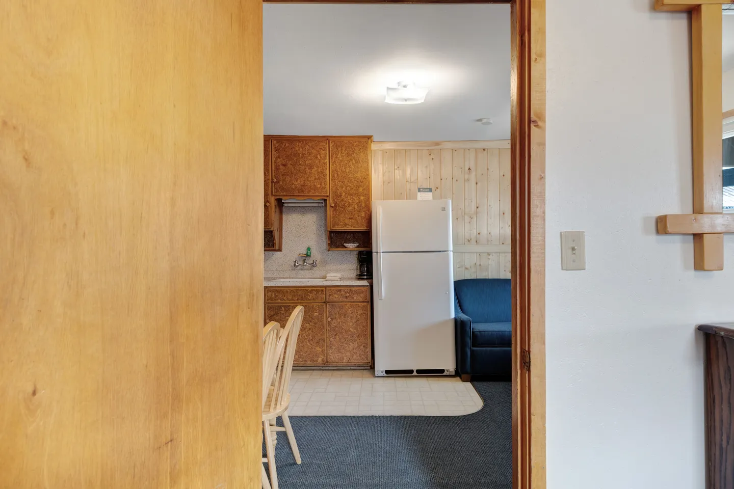 A view through an open doorway looking into a kitchen area with wooden cabinets, a white refrigerator, and a small adjoining seating space.