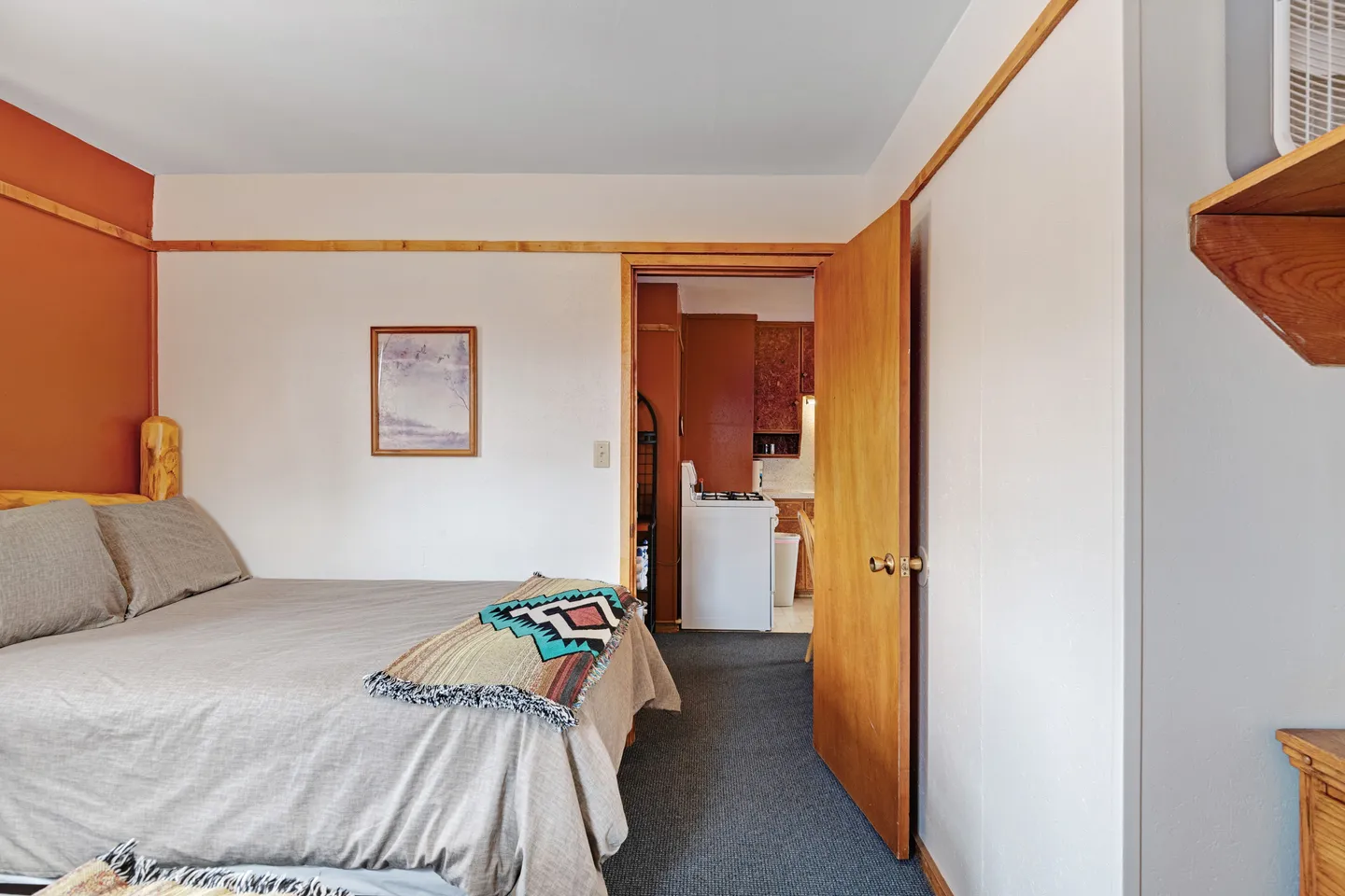 Bedroom with a bed dressed in neutral linens and an open wooden door leading toward a kitchen area.