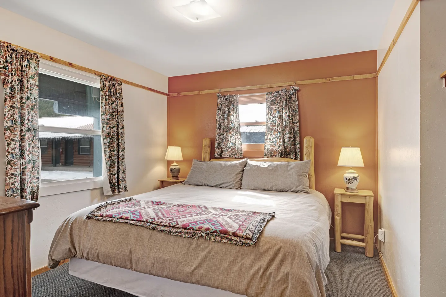 Bedroom with a bed made up with a neutral duvet, rustic wooden headboard, small wooden nightstands with lamps, and two windows with patterned curtains.