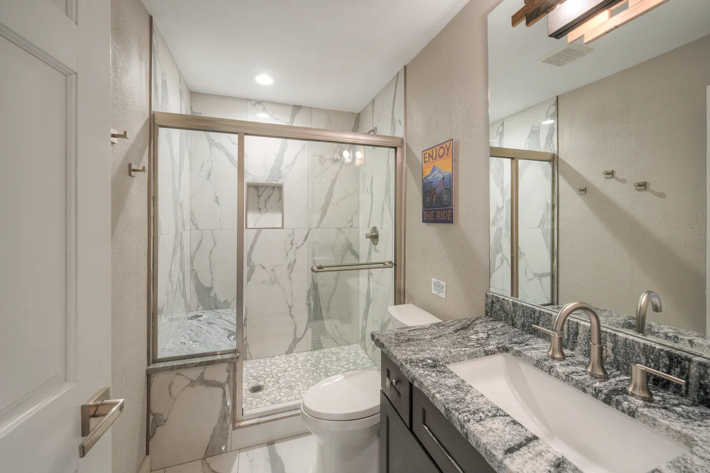 Bathroom showing a vanity with dark cabinets and a mottled stone countertop beside a toilet and a walk-in shower with sliding glass doors and marble-patterned tile.