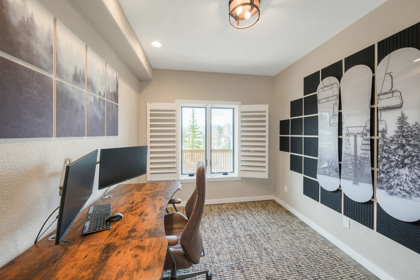 Dedicated home office space with a large curved wooden desk, two computer monitors, an office chair, and ski-lift themed wall art.