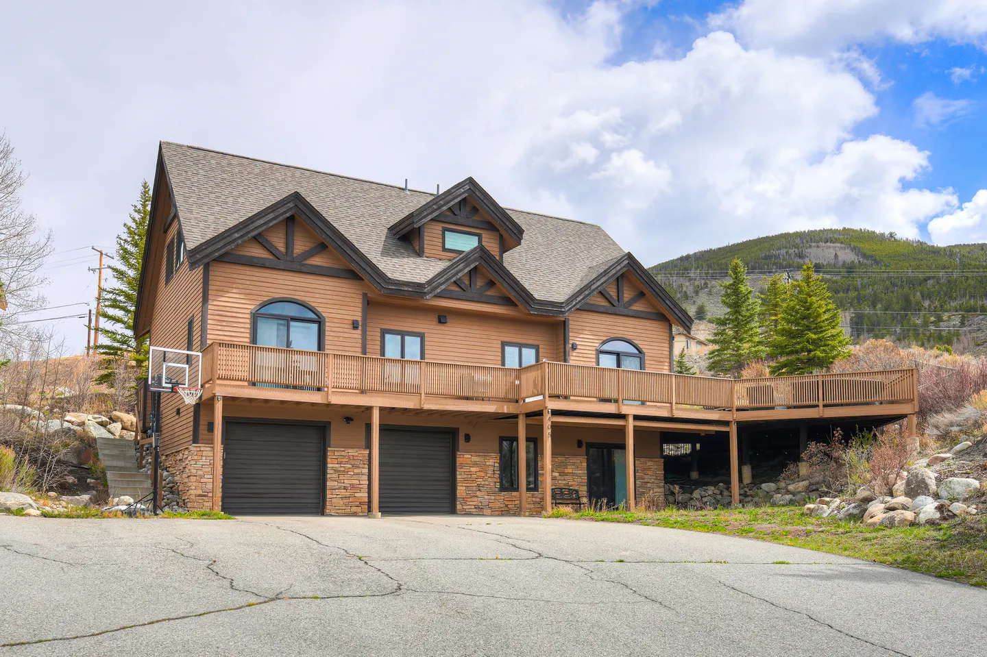 Exterior view of a multi-level wood-sided home with a stone-accented ground floor, elevated wraparound deck, and paved driveway.