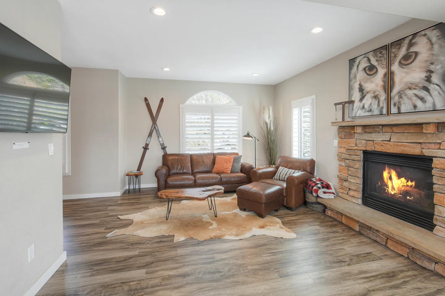 Living room with brown leather seating, a stone-faced fireplace with lit fire, wood-look flooring, and a wall-mounted TV.