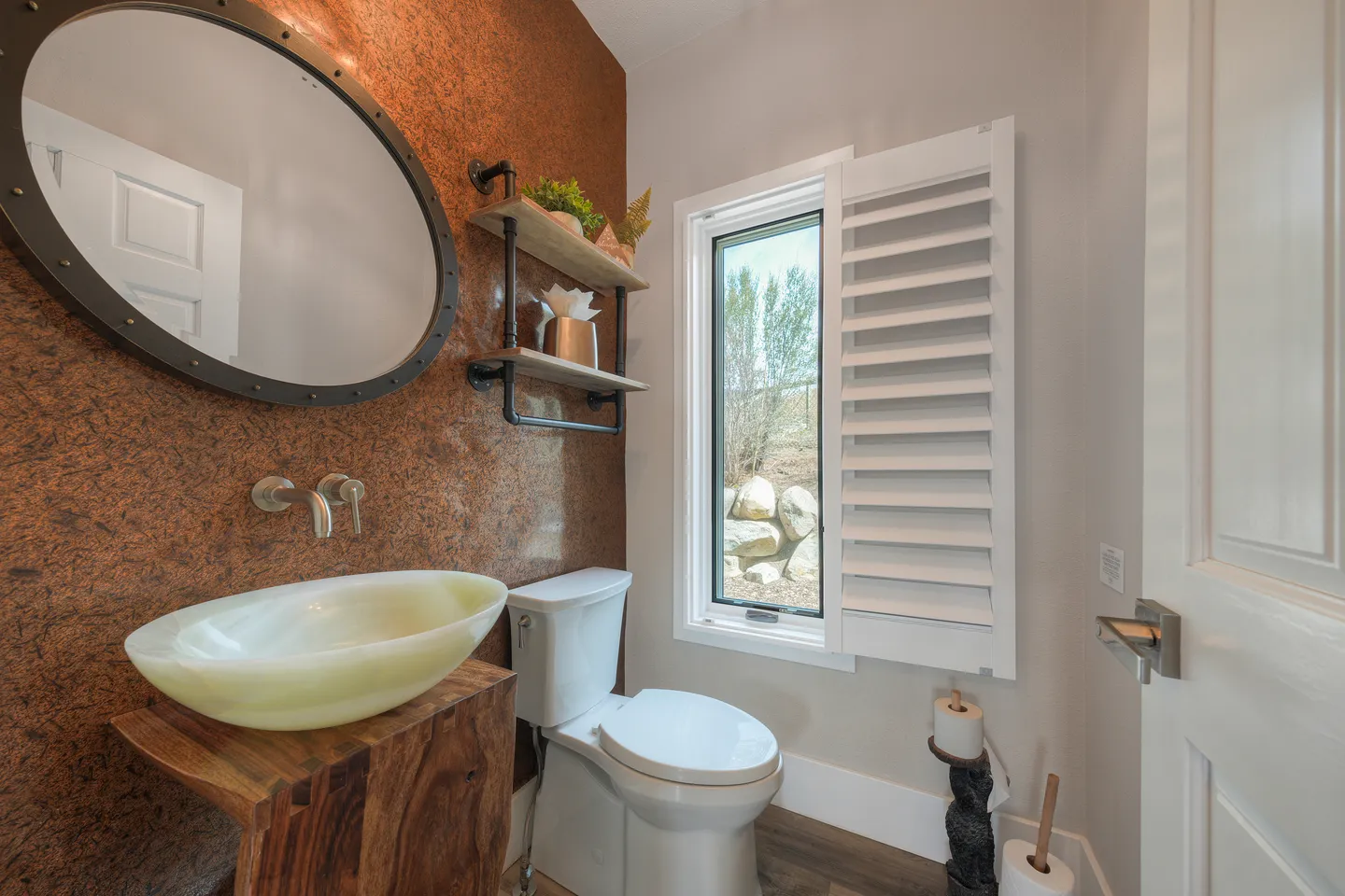 Bathroom with a toilet, vessel sink on a wooden stand, round mirror with rivets, textured copper-toned accent wall, and a window with white shutters.