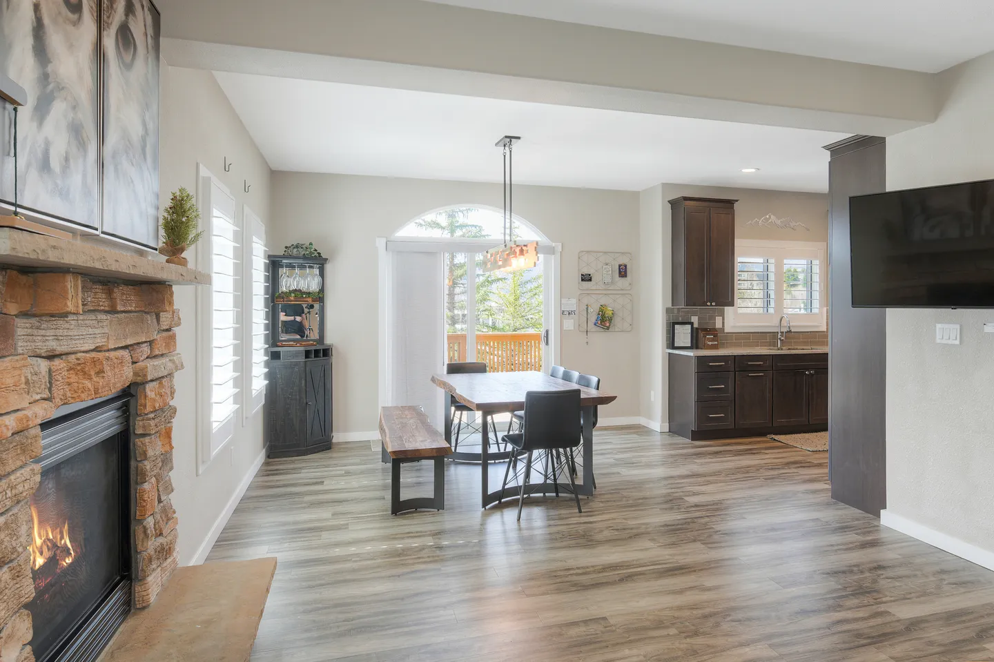 Open-concept living and dining area featuring a stone fireplace with an active fire, wood dining table with chairs and bench, and a view into the kitchen.