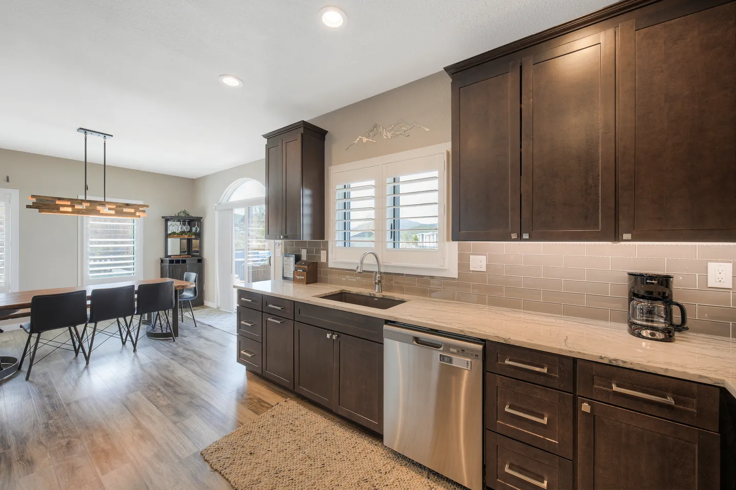 Kitchen counter with dark wood cabinetry, stainless steel dishwasher, undermount sink, and adjacent dining area with a table and chairs.