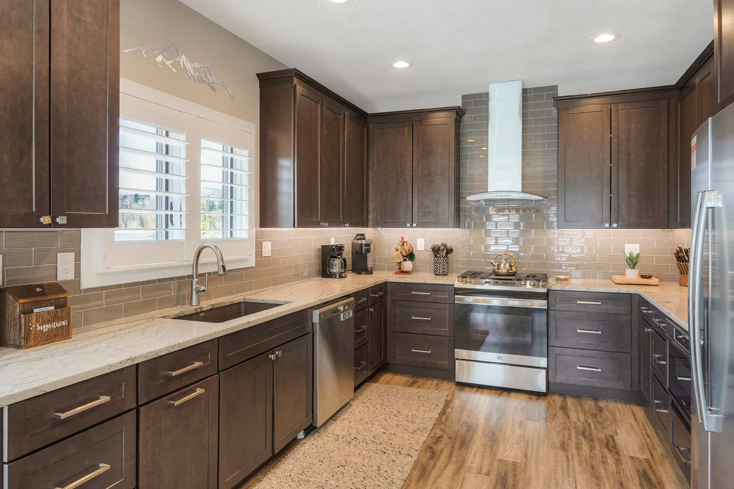 A kitchen with dark wood cabinetry, light countertops, stainless steel appliances, and a window with white shutters.