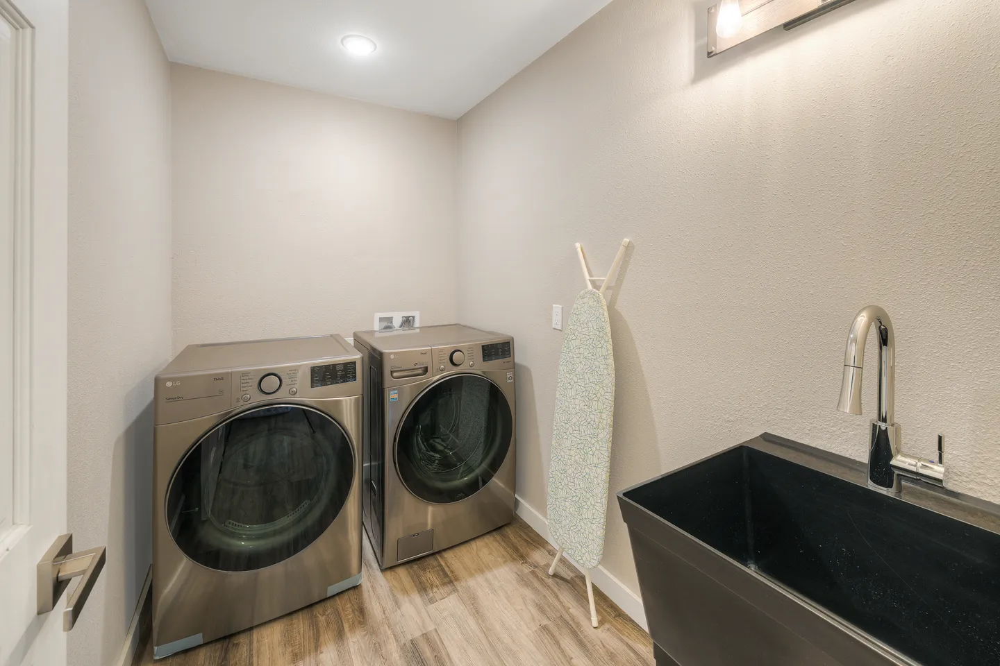 A laundry room with a metallic front-loading washer and dryer, an ironing board, and a utility sink.