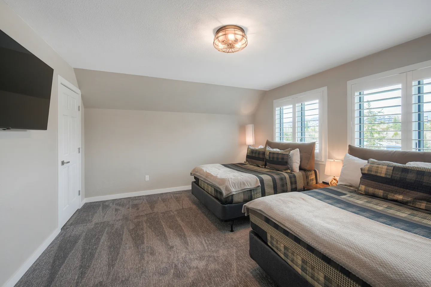 Bedroom with two beds, plaid bedding, gray carpeting, shuttered windows, and a wall-mounted TV.