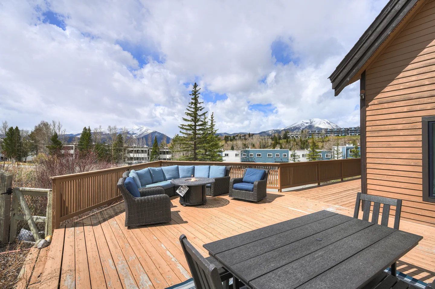 Spacious outdoor wooden deck with sectional patio seating, a fire pit table, and a dining set overlooking distant mountains.