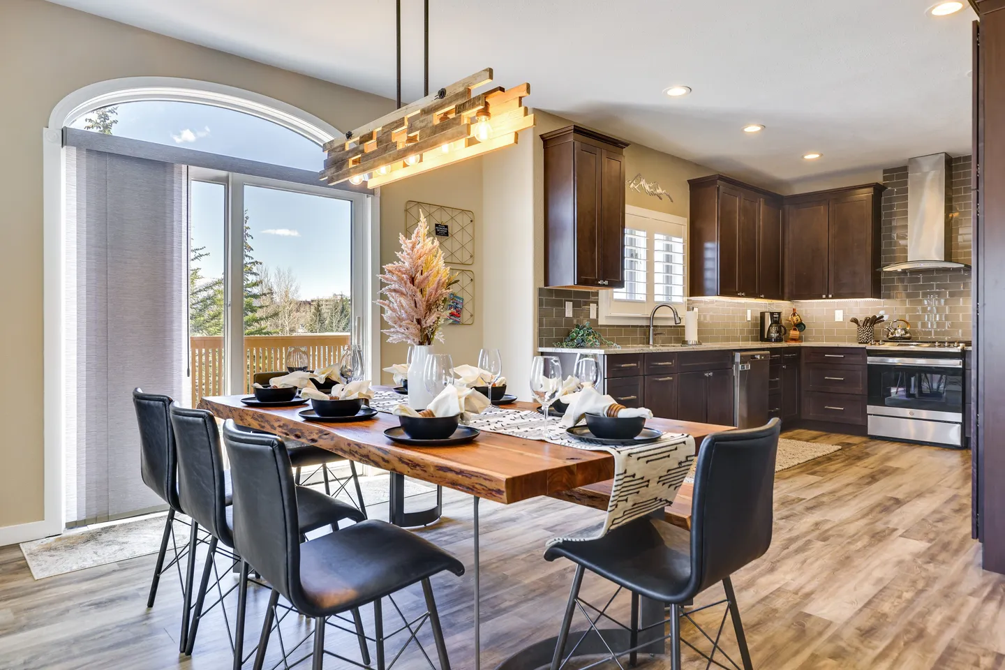 Dining area with a live-edge wooden table and black chairs set for a meal, situated next to an arched glass door leading to a balcony and an open-concept kitchen.