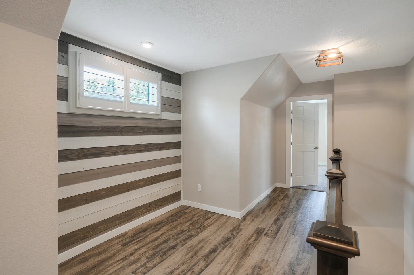 Upstairs landing with wood-look flooring, a dark banister, an open doorway, and an accent wall with multi-toned horizontal wood planks beneath a shuttered window.
