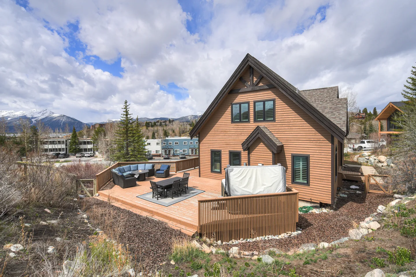 Exterior view of a brown two-story house with a wooden deck featuring a dining set, sectional sofa, and covered hot tub with mountains in the background.