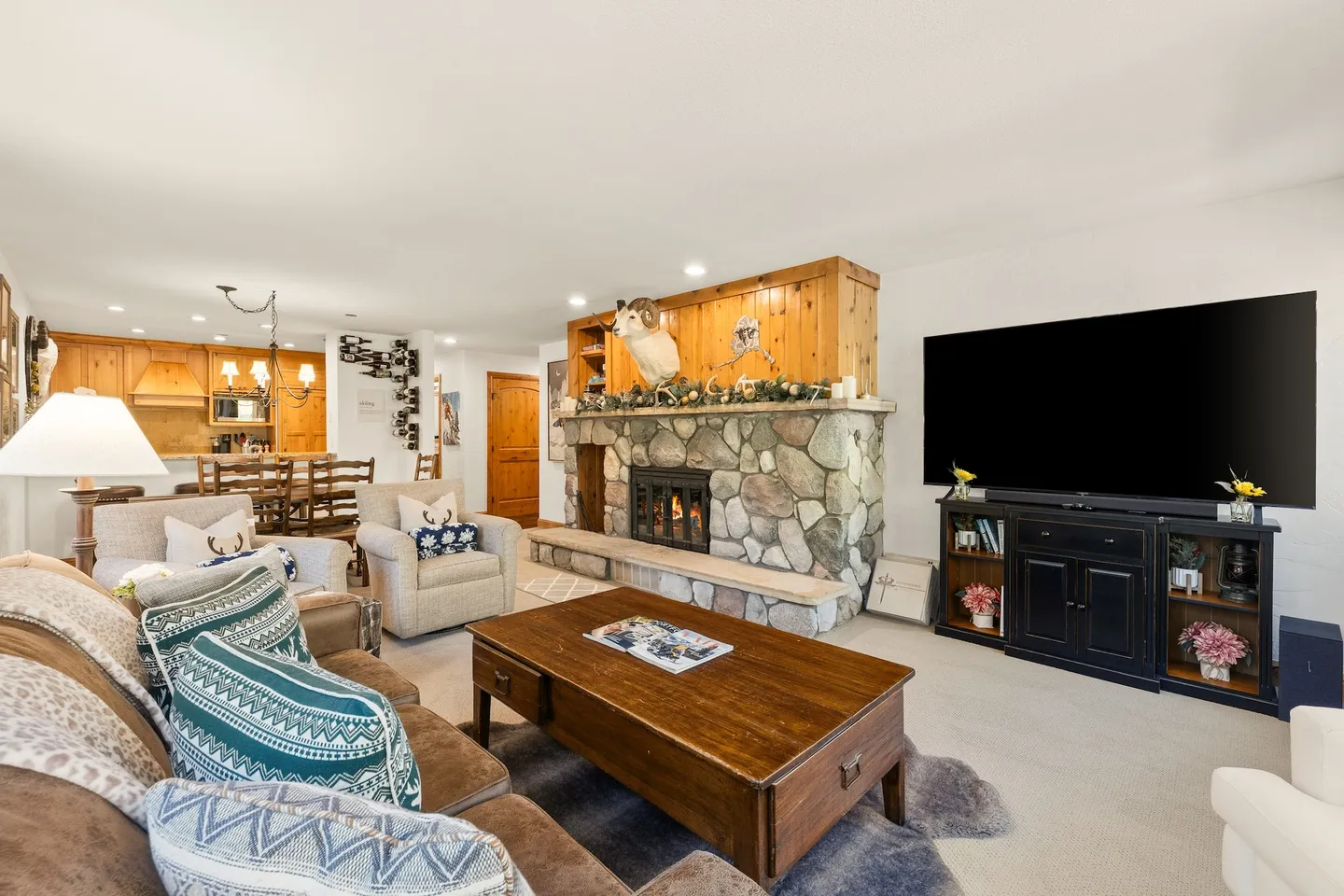 Open-concept living room featuring a stone fireplace, large television, armchairs, and adjacent dining area.