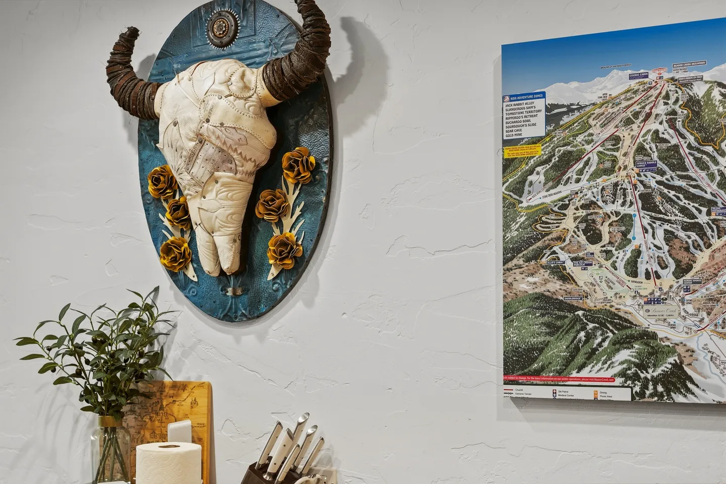 Decorative bull skull art piece with yellow roses mounted on a white textured wall alongside a ski resort trail map.